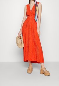 Rotes, plissiertes Maxikleid mit V-Ausschnitt und dünnen Trägern. Kombiniert mit einer Strohtasche und sandfarbenen Plateau-Sandalen. Bunter Schal um den Hals gebunden.