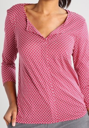 Vrouw draagt een roze blouse met lange mouwen, kleine witte geometrische patronen en een ronde kraag, gecombineerd met grijze broek.