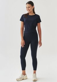 Navy sinine aktiivsusriiete komplekt, mis sisaldab sobivat lühikeste varrukatega toppi ja kõrge vöökohaga retuuse, mõlemad sujuvast ja elastse kangaga. Sneakerid on beežid.