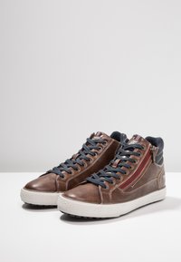 Brown leather high-top sneakers with navy laces, a red side zipper, and textured accents. White rubber sole with tread pattern.