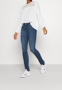 Vit långärmad blus med spetsdetaljer, tillsammans med mörkblå slim fit-jeans och vita sneakers, som fångar en avslappnad outfit.