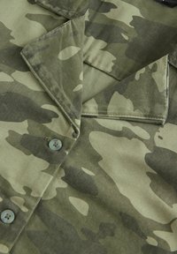 Zelená maskovací košile s klopovým límcem, zapínáním na knoflíky a variacemi v odstínech barev. Hladká textura látky.