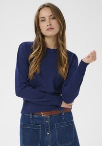 Pull en tricot bleu marine à manches longues avec poignets côtelés, présentant des détails bouffants aux épaules. Porté avec une jupe en denim.