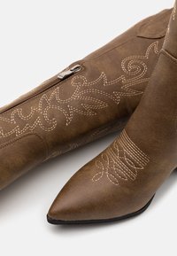 Hnědé kožené kotníkové boty s špičatou špičkou, které mají složité vzory prošívání a boční zip pro zapínání.