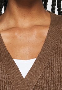 Cardigan côtelé marron avec un col en V profond, superposé sur un haut blanc ajusté. Accent sur la texture et le contraste des couleurs.