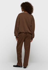 Persona con cabello rizado que lleva un suéter marrón holgado y pantalones anchos a juego, de pie de espaldas sobre un fondo liso.