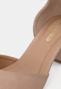 Chaussure à talon haut beige en daim à bout ouvert avec une semelle intérieure rembourrée et un talon bloc, inclinée pour montrer le marquage intérieur.