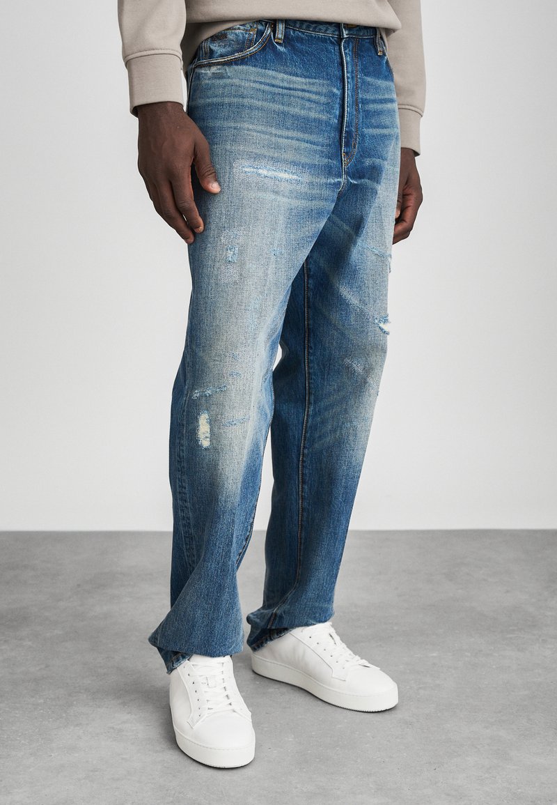 Jeans de mezclilla azul desgastados con parches descoloridos y bordes deshilachados, que presentan un corte relajado. Combinados con zapatillas altas blancas.