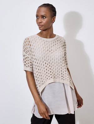 Maglione lavorato a maglia color beige chiaro con un design a trama aperta, maniche corte e un effetto a strati sopra una camicetta grigia chiara, abbinato a pantaloni neri.