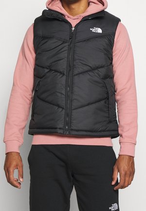 Mann trägt eine schwarze ärmellose Steppweste über einem rosa Hoodie und schwarze Hose mit sichtbaren The North Face-Logos auf Weste und Hose.