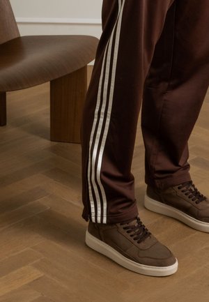 Braune Sporthosen mit drei vertikalen weißen Streifen an den Seiten. Braune Sneakers mit weißen Sohlen, strukturierten Schnürsenkeln und einer glatten Oberfläche.