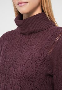 Strikket turtleneck-genser i dyp lilla med et teksturert bladmønster og ribbet krage. Har åpne strikkedetaljer på skulderen.