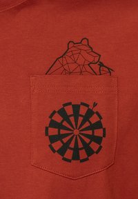 Camiseta de color óxido con un gráfico geométrico de un oso negro encima de un bolsillo, y un diseño circular de un tablero de dardos negro en el bolsillo.