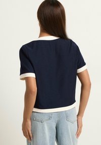 Mujer con pelo largo y castaño que lleva una camiseta de manga corta azul marino con ribete blanco y jeans de mezclilla azul claro, vista desde atrás.