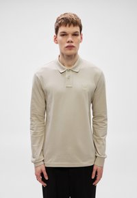 Bež polo majica z dolgimi rokavi, ovratnikom, dvema gumboma in majhnim kvadratnim logotipom na levi strani prsi. Gladek material in oprijeta oblika.