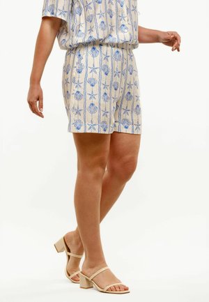 Kvinde iført beige shorts med blå muslinge- og søstjerneprint og beige sandaler med lav hæl, stående på en hvid baggrund.