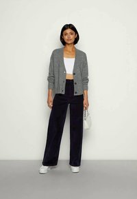 Mulher vestindo um cardigan cinza com botões, uma t-shirt curta branca, calças largas pretas, tênis brancos, segurando uma pequena bolsa branca, de pé contra um fundo liso.
