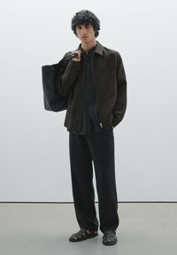 Veste en laine noire avec fermeture éclair, chemise à boutons, large pantalon noir et sandales à lanières. Le mannequin tient un grand sac fourre-tout noir.