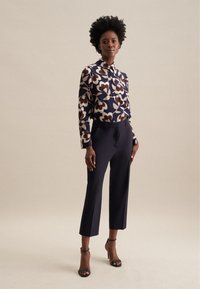 Blusa floreale in navy, crema e bordeaux con collare e dettagli dei bottoni, abbinata a pantaloni neri aderenti e tacchi a spillo neri.
