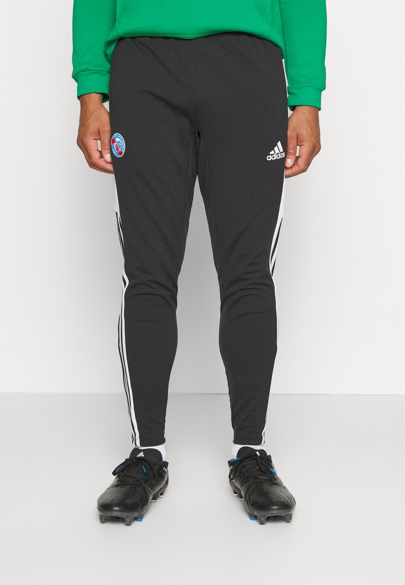 Schwarze, schmal geschnittene Sporthosen mit weißen Seitenstreifen und einem runden Logo auf dem Oberschenkel. Kombiniert mit schwarzen Fußballschuhen.