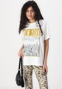 Bela kratka majica s kratkimi rokavi, ki prikazuje napis "NEW YORK CITY OF DREAMS" in sivo grafiko mestnega obzorja. V kompletu z teksturiranimi hlačami z vzorcem kače.