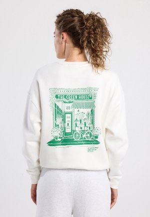 Felpa bianca con una grafica verde che rappresenta un negozio etichettato "THE GREEN HOUSE." Le caratteristiche includono una bicicletta e piante in vaso.