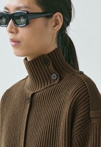 Brauner gerippter Strickpullover mit hohem Kragen und Knopfakzenten am Hals. Die Textur ist weich und das Design ist strukturiert.