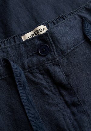 Granatowe lniane spodnie z zapięciem na guziki, elastycznym paskiem w talii, sznurkiem do regulacji i częściowo widoczną białą metką.