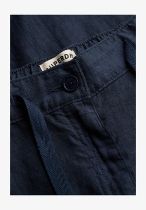 Granatowe lniane spodnie z zapięciem na guziki, elastycznym paskiem w talii, sznurkiem do regulacji i częściowo widoczną białą metką.