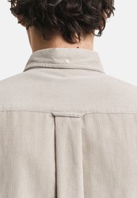 Achteraanzicht van een persoon die een beige corduroy shirt draagt met een button-down kraag en geplooide details op de bovenrug.