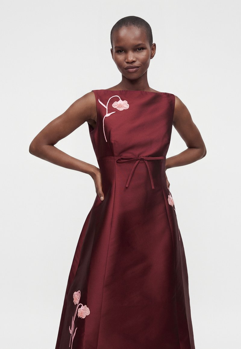Femme debout, mains sur les hanches, portant une robe bordeaux sans manches ornée de broderies florales roses et d'une ceinture nouée à la taille.