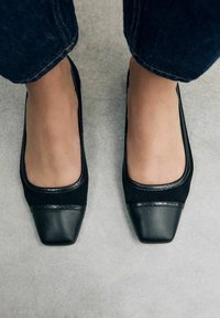 Ballerine nere in suede con una punta quadrata e una punta in pelle nera lucida. Il bordo texturizzato lungo il perimetro aggiunge dettaglio.