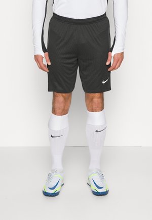 Person iført sorte Nike sportsshorts, hvid langærmet trøje, hvide Nike knæstrømper og hvide sportssko med blå og neon accenter.