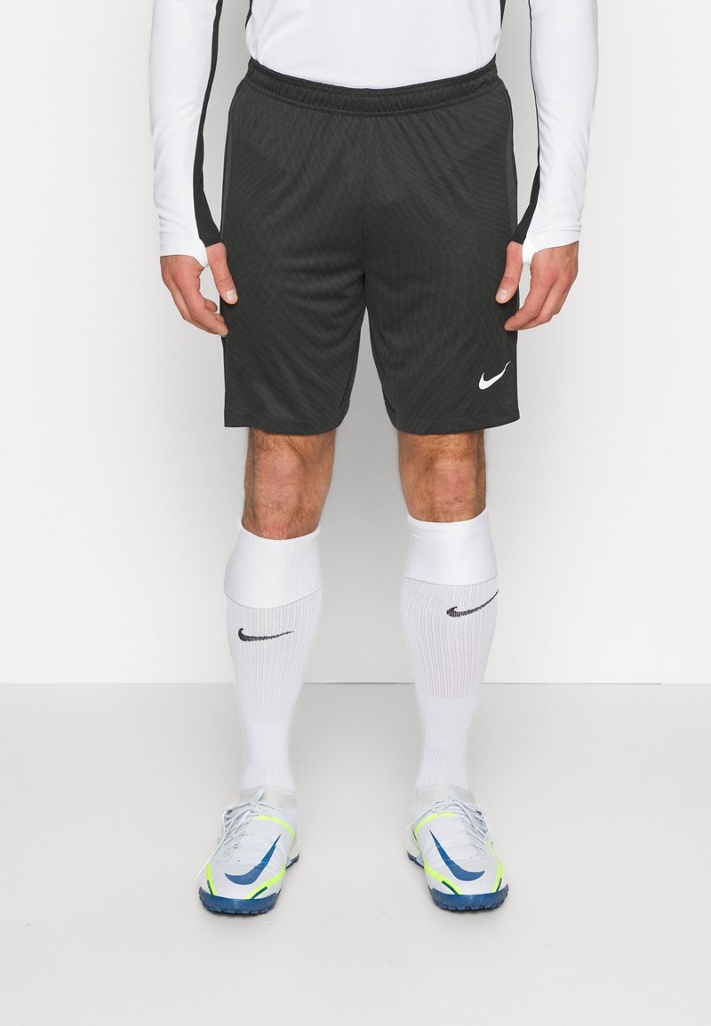 Personne portant un short de sport Nike noir, un haut à manches longues blanc, des chaussettes Nike blanches montantes jusqu'aux genoux et des chaussures de sport blanches avec des touches bleues et néon.