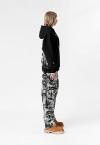 Sudadera negra de gran tamaño con cordones, combinada con pantalones de patrón blanco y negro. Calzado marrón con suela gruesa.