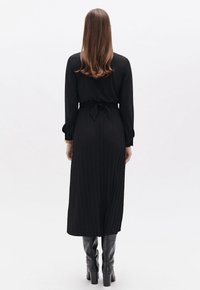 Schwarzes Langarmkleid mit plissee Art Rock und Taillengürtel, kombiniert mit kniehohen schwarzen Stiefeln. Glatter Stoff und maßgeschneiderte Gestaltung.