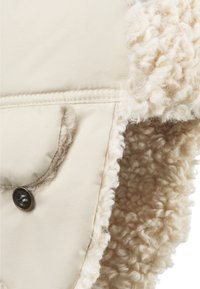 Beige stofbeklædning med en fluffy, cremefarvet fleece-fòr. Har en knaplukket lomme og teksturerede, bløde kanter.