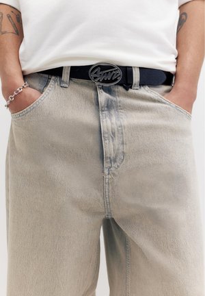 Osoba s rukami v vreckách nosí svetlo vypraté rifle, bielu košeľu, čierny opasok s oválnou prackou, strieborný náramok a viditeľné tetovania na rukách.