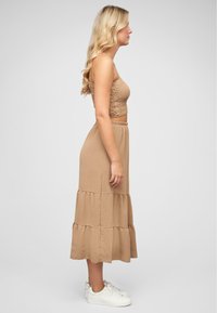 Beige twee-delige outfit met een smock tube top en een gelaagde maxi rok. De stof is zacht met een lichte textuur, gecombineerd met witte sneakers.