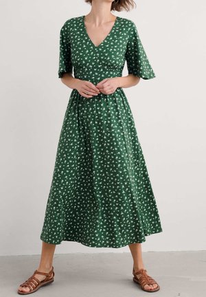 Femme en robe midi verte à imprimé floral blanc, col en V, manches courtes, debout les mains jointes, portant des sandales marron.