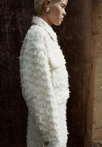 Witte textuurjack met rafelige details, met een kraag en een voorzak. Ggedragen met grote ronde oorbellen, getoond vanuit een zijaanzicht.