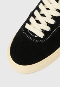 Melnas zamšādas sporta apavi ar krēmkrāsas šņorēm, noapaļotu purngalu un kontrastējošu bēša gumijas zolīti. Redzamas šuvju detaļas.