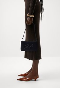 Marin läderhandväska med präglad "Karl"-logotyp, matchad med en brun ribbad klänning och orange lackade pumps med spetsiga tår. Guld kedjearmband som accent.