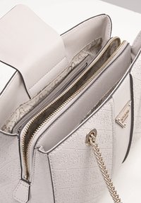 Sac à main gris clair en matériau texturé, avec un compartiment zippé, du matériel doré, et une bandoulière en chaîne. Accent logo notable à l'avant.