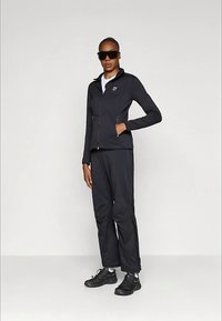 Kvinne iført svart jakke med glidelås, matchende svarte bukser, svarte sneakers, hvite sokker og store svarte solbriller, som står mot en enkel bakgrunn.