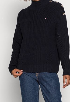Person iført en sort ribstrikket sweater med knapper på den ene skulder og blå denim jeans, holder nederdelen af sweateren med den ene hånd.