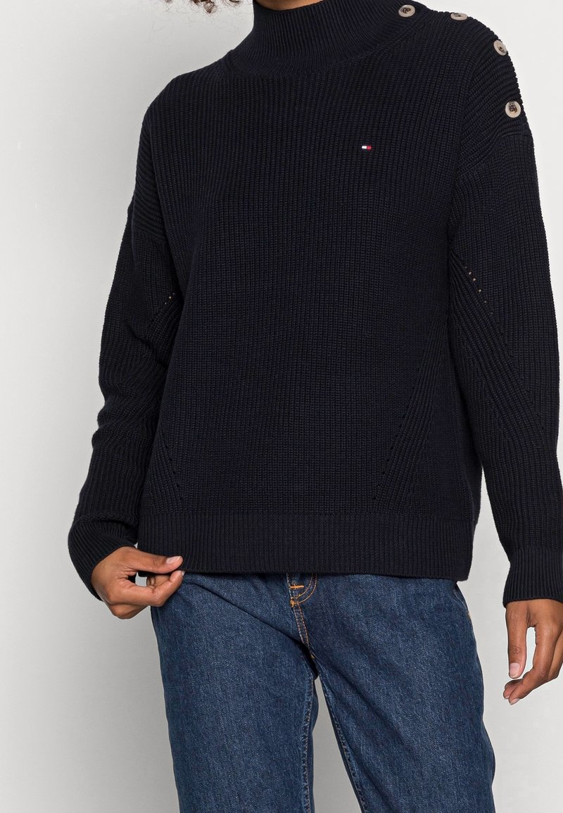 Personne portant un pull en maille côtelée noire avec des boutons sur une épaule et un jean bleu en denim, tenant l’ourlet du pull d’une main.