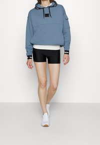 Sweat-shirt bleu surdimensionné avec capuche, logo noir et cordons ; associé à un short de sport noir et des baskets blanches.