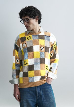 Homme aux cheveux bouclés et lunettes, portant un pull multicolore à carreaux avec des visages d'ours et des motifs souriants, une main dans la poche de son jean.