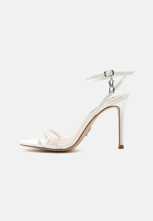 Λευκό σανδάλι με ψηλό τακούνι, λεπτά λουράκια, αγκράφα στον αστράγαλο και μεταλλική λεπτομέρεια σε ουδέτερο φόντο.